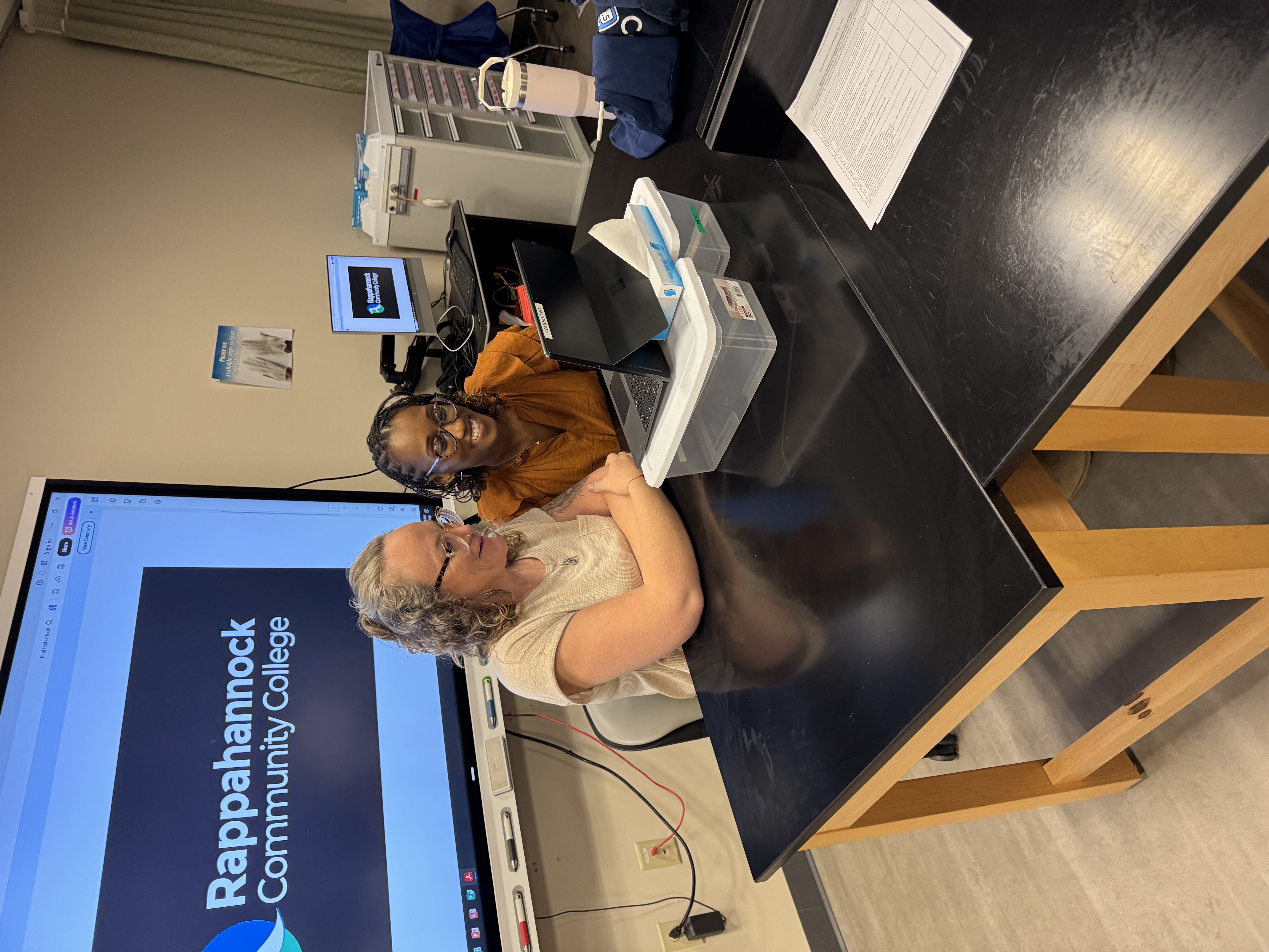 Two women sit at a classroom lab table working together on a laptop, with a screen behind them displaying “Rappahannock Community College.”