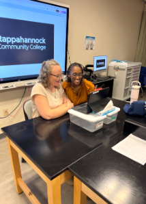 Two women sit at a classroom lab table working together on a laptop, with a screen behind them displaying “Rappahannock Community College.”