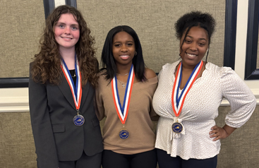 Three women stand smiling indoors, each wearing a medal on a red, white, and blue ribbon.