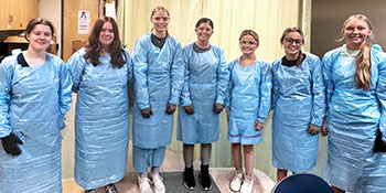 Group of students and an instructor wearing blue disposable gowns stand in a lab classroom.